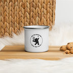 White enamel mug with a black bear graphic and text on a wooden surface with cookies and a woven basket in the background.